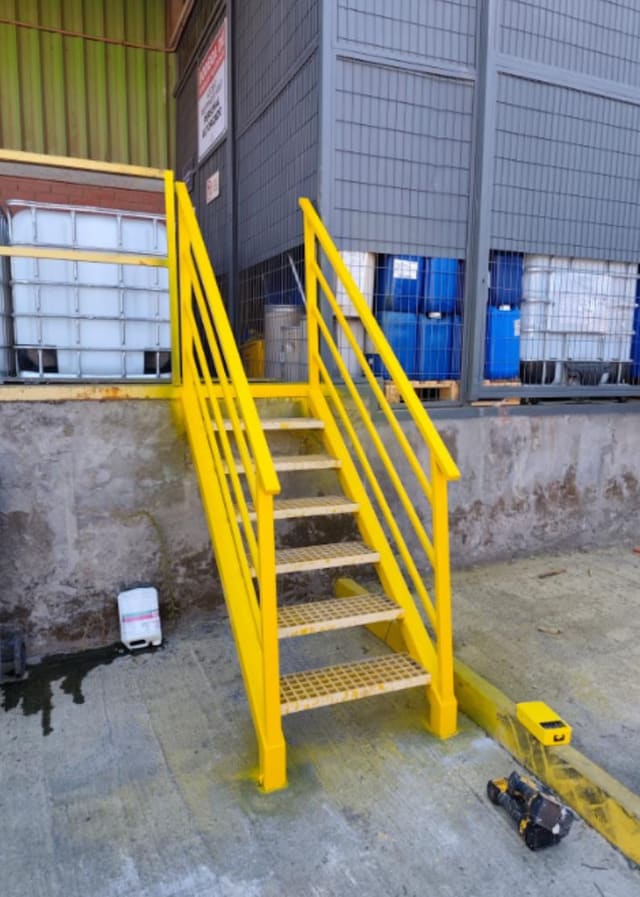 Escalera metálica pintada de amarillo con barandas y peldaños de rejilla antideslizante en el exterior de una planta