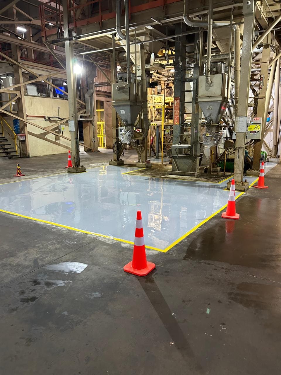 Piso epóxico gris recién aplicado en una planta industrial, con demarcación amarilla y conos de seguridad